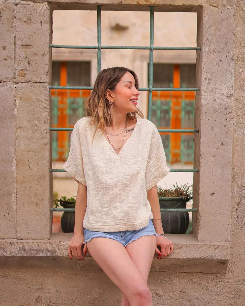 Woman in a Beige double gauze cotton v neck top and denim shorts sitting on a stone ledge with a window in the background.