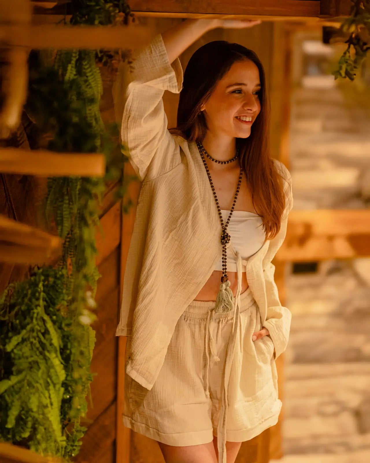 Woman in a beige cotton loungewear set outfit standing next to a wooden structure with greenery
