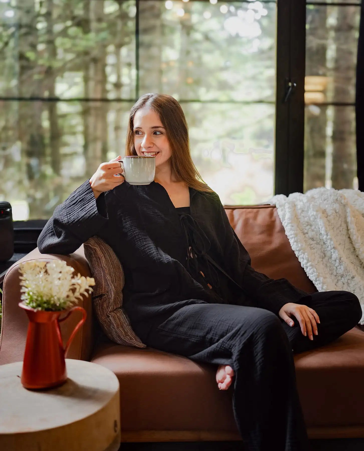 A Black woman in black loungewear set is seated on a couch, holding a cup, with a backdrop of trees visible through the window