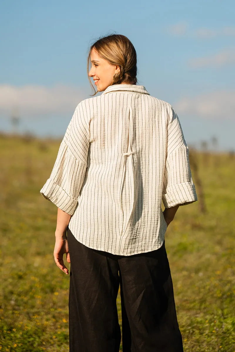 Woman wearing a white colored striped shirt and dark pants standing in a field with a clear sky.