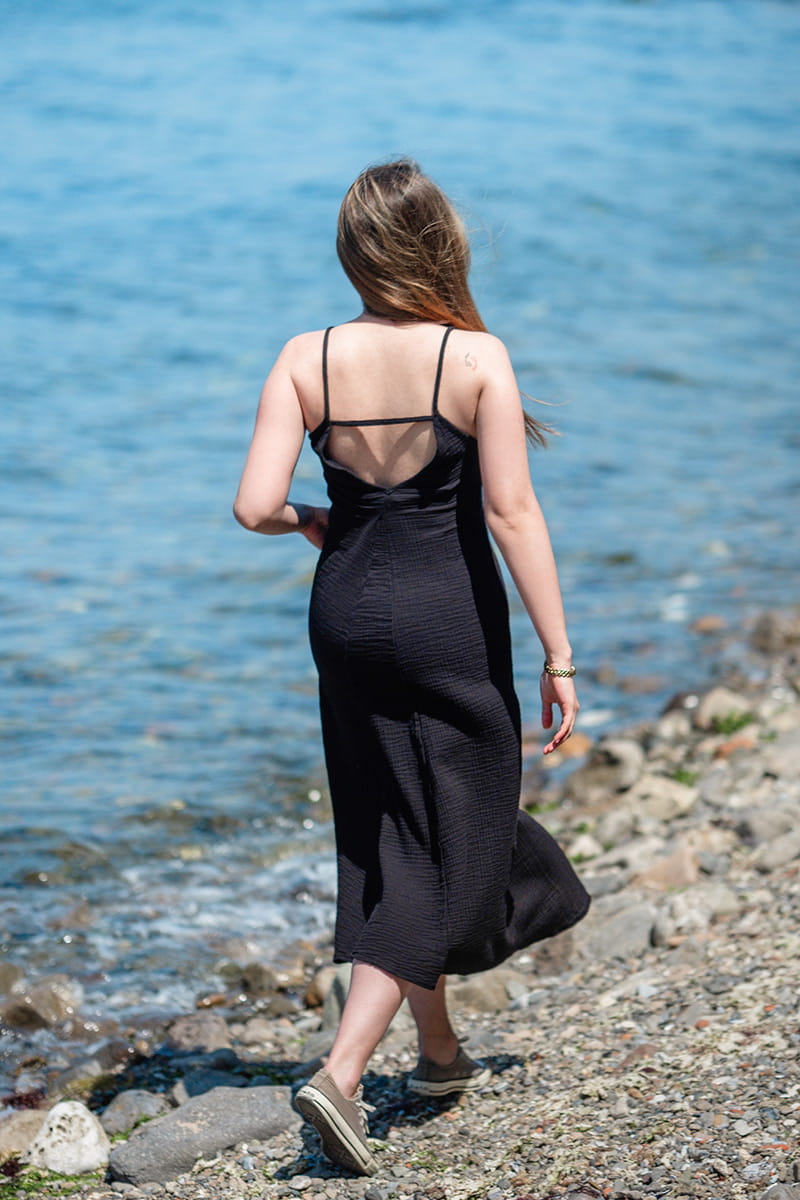 Woman in a black Double Gauze Cotton  Dress walking along a rocky shoreline with blue water in the background