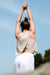 A woman in a cross-back tank top stands with arms lifted against a bright blue sky