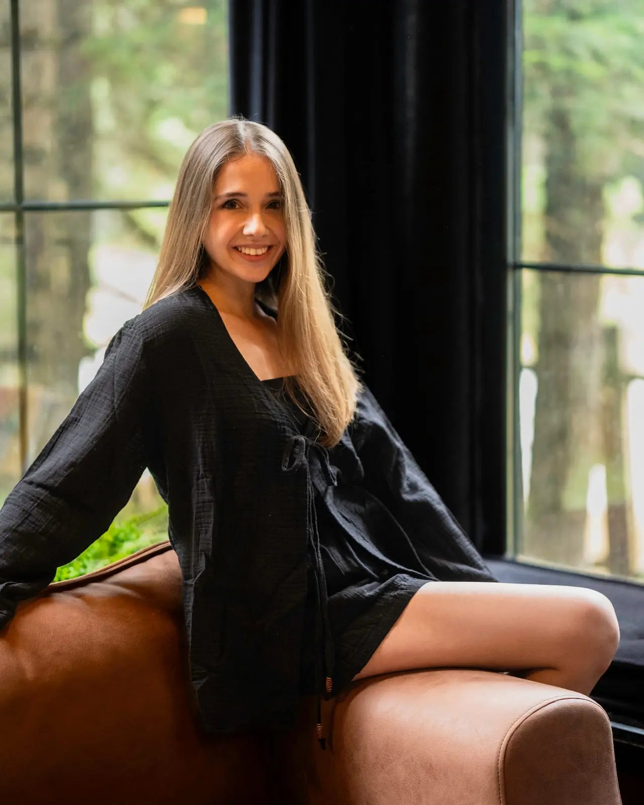 Woman in a black lounge wear sitting on a brown leather chair with large windows in the background
