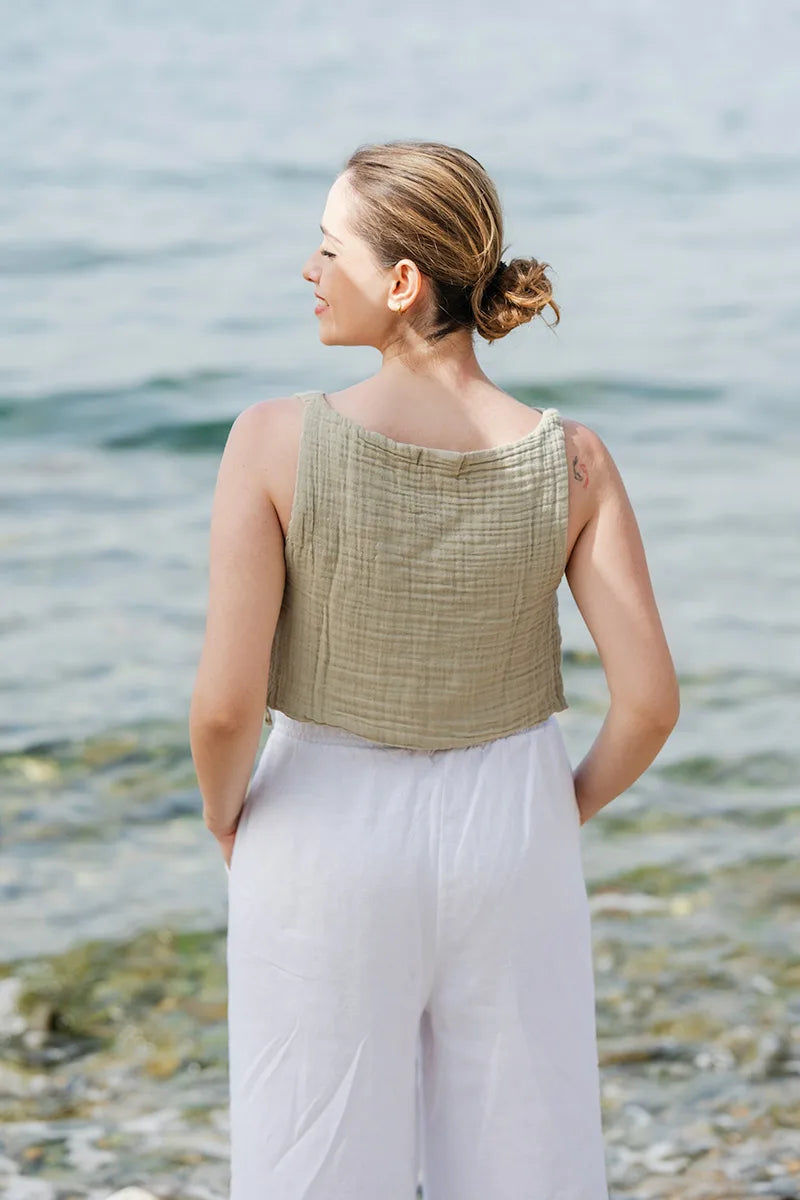 Woman standing by the water wearing a green double gauze top and white linen pants.