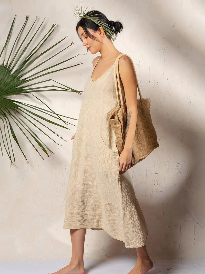Woman in a beige double gauze dress holding a brown tote bag against a white wall with a plant.