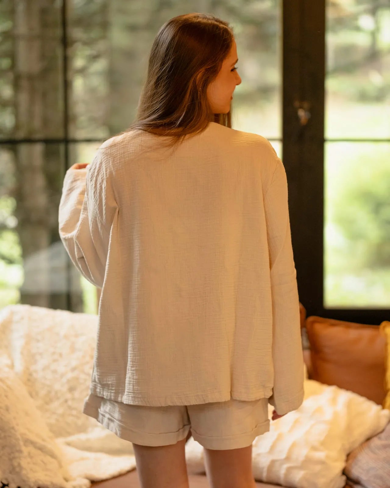 Woman wearing a beige cotton pyjama set standing in a room with large windows.