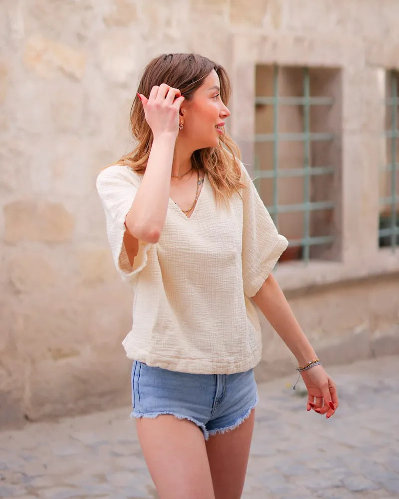 Woman wearing a beige-colored blouse top and denim shorts standing against a stone wall.