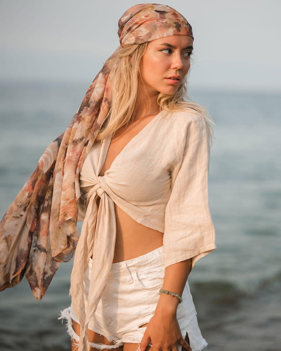 Woman on a beach wearing a beige top and white shorts with a patterned headscarf.