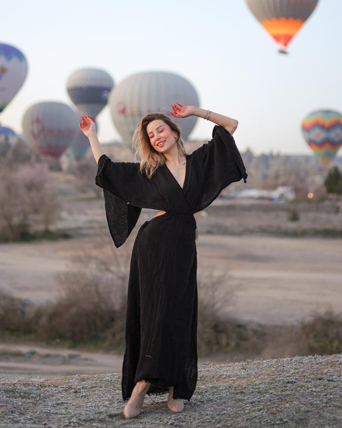 A woman in a black v-neck maxi dress standing with her arms raised, in front of hot air balloons in a field.