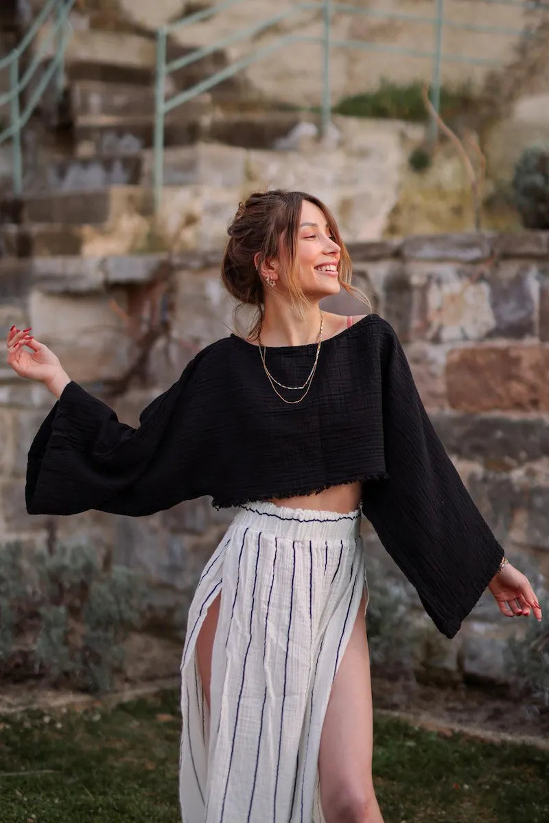 Woman in black off the shoulder top and white striped skirt standing outdoors with stone steps and greenery in the background