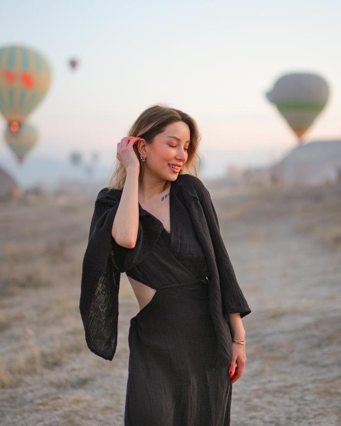 A woman in a black V-neck dress stands smiling in front of colorful hot air balloons against a clear blue sky.