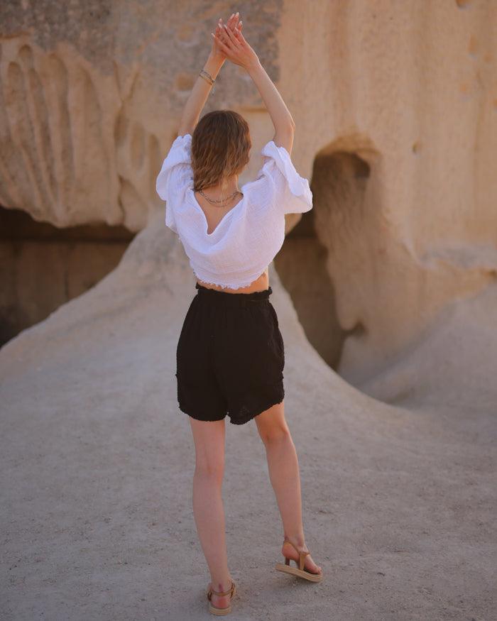 A woman in a white shirt and black crinkle cotton shorts stands confidently in front of a large rock formation.