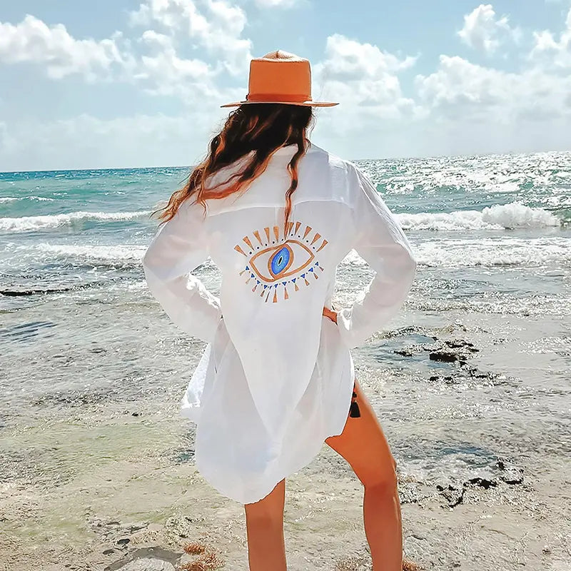Person wearing a white shirt dress with a blue eye design on a beach.