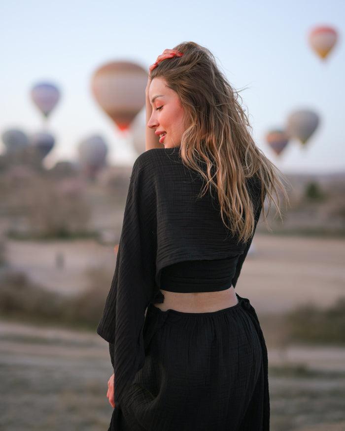 A woman in an open-back black cotton dress stands before colorful hot air balloons against a clear sky.