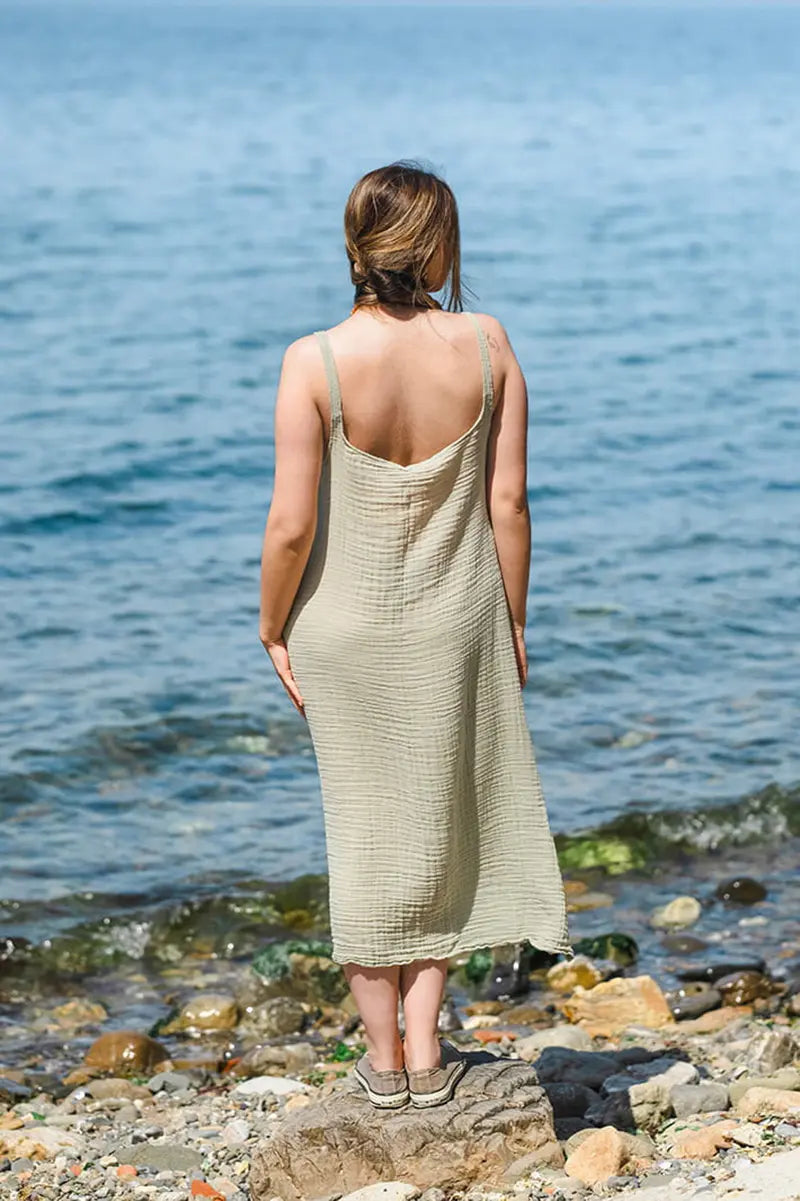 Woman in a green midi dress standing on a rocky beach looking at the ocean.