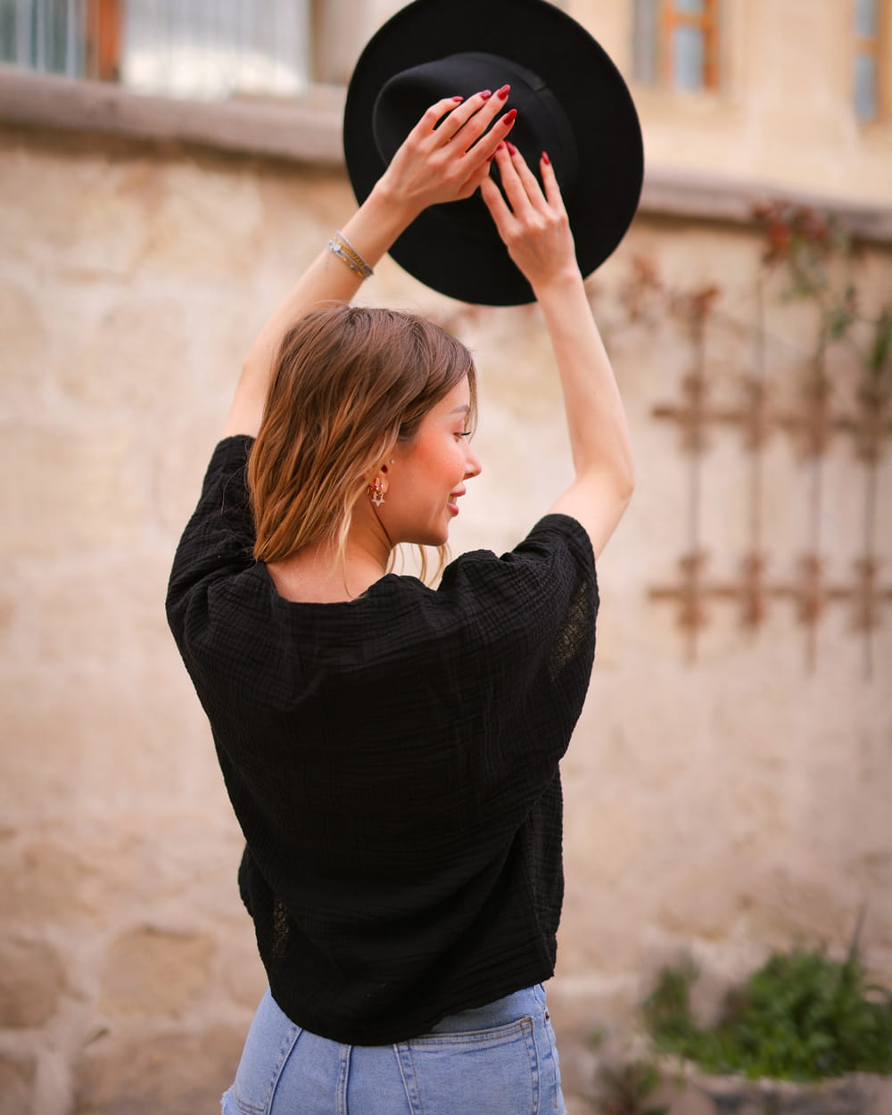 A woman dressed in a black short-sleeve blouse made of crinkle cotton is holding a black hat over her head in front of a stone wall
