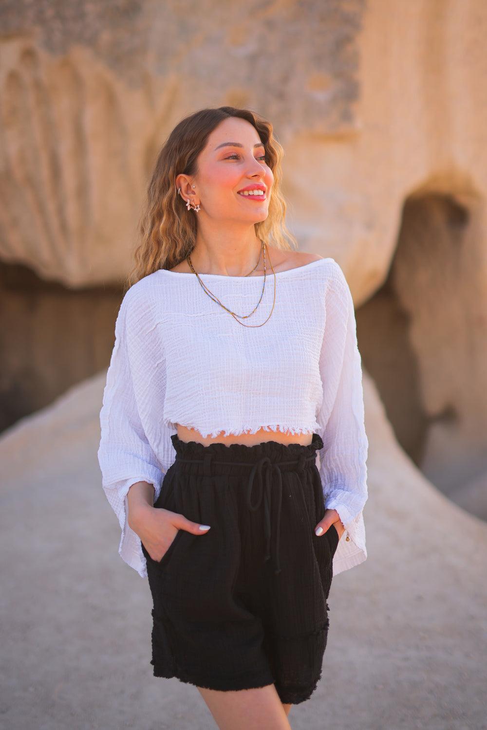 A woman standing outdoors wearing a white off shoulder crop top and black shorts.