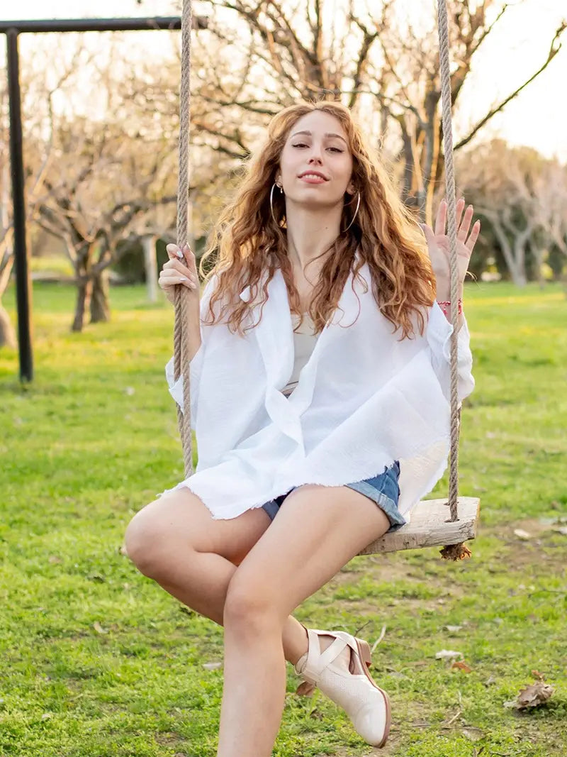 Woman in a white 4-layer-muslin-kimono and denim shorts sitting on a swing in a park.