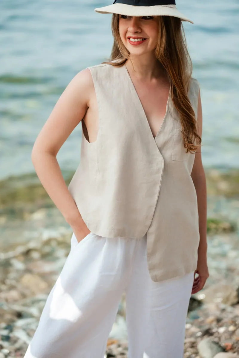 Woman wearing a beige sleeveless linen vest and white pants standing by a body of water.