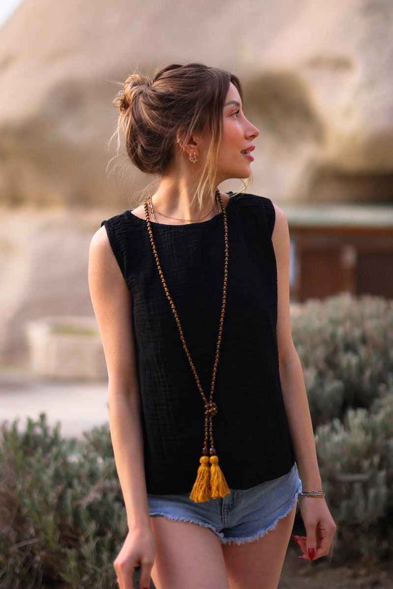 Woman wearing a black sleeveless top and denim shorts with a long necklace, standing outdoors.