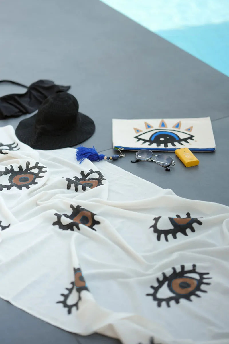 White beach towel with eye designs, black hat, sunglasses, and pouch on a gray surface.