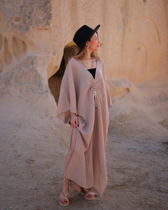 A woman in a long muslin dress and hat stands gracefully in front of a striking rock formation.