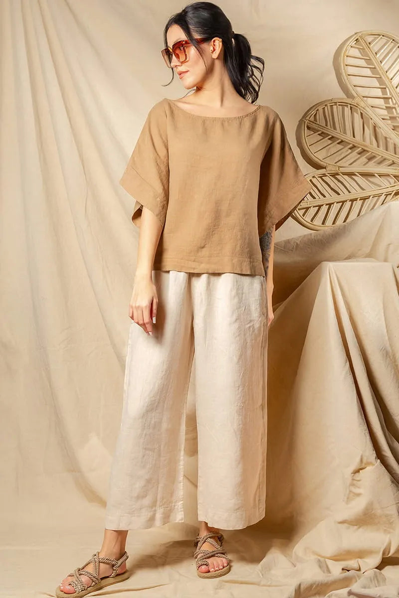 Woman wearing a beige top and  Beige Linen Palazzo Pants standing against a beige curtain with a decorative leaf.