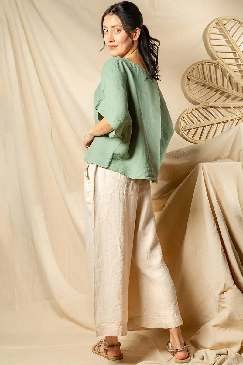 Woman wearing a green top and beige linen pants standing against a beige curtain with a decorative leaf.