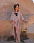 A woman wearing a beige gauze dress with long sleeves, styled in a kaftan style. She is standing in front of large rocks, wearing a wide-brimmed hat and platform shoes.