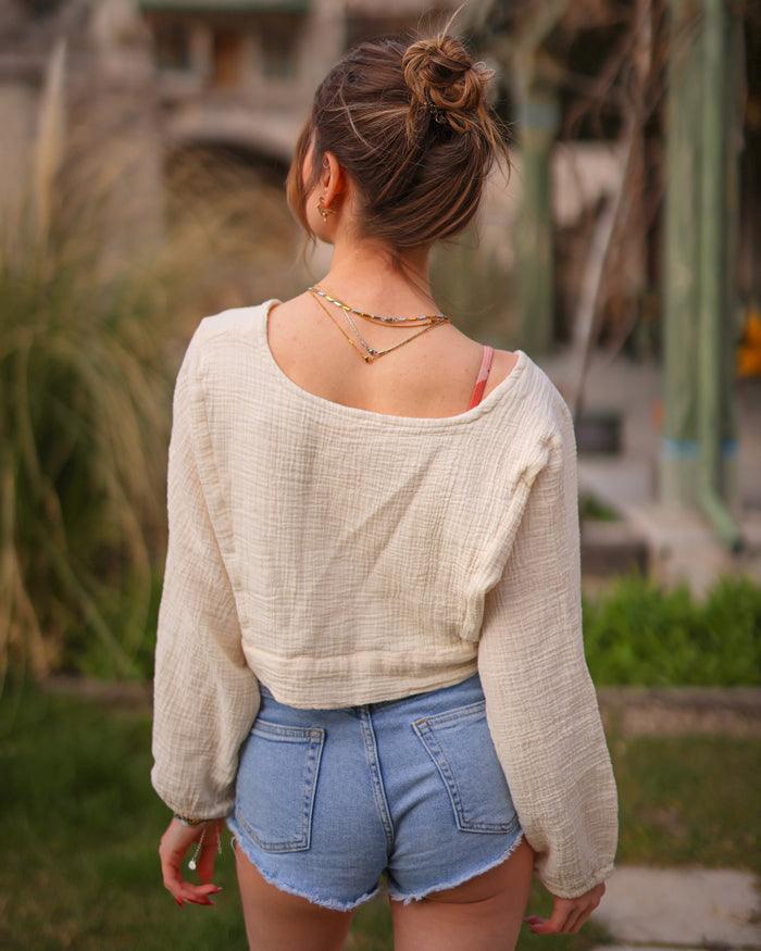 A woman wearing a beige double gauze cotton crop top and denim shorts stands casually, enjoying a sunny day outdoors.