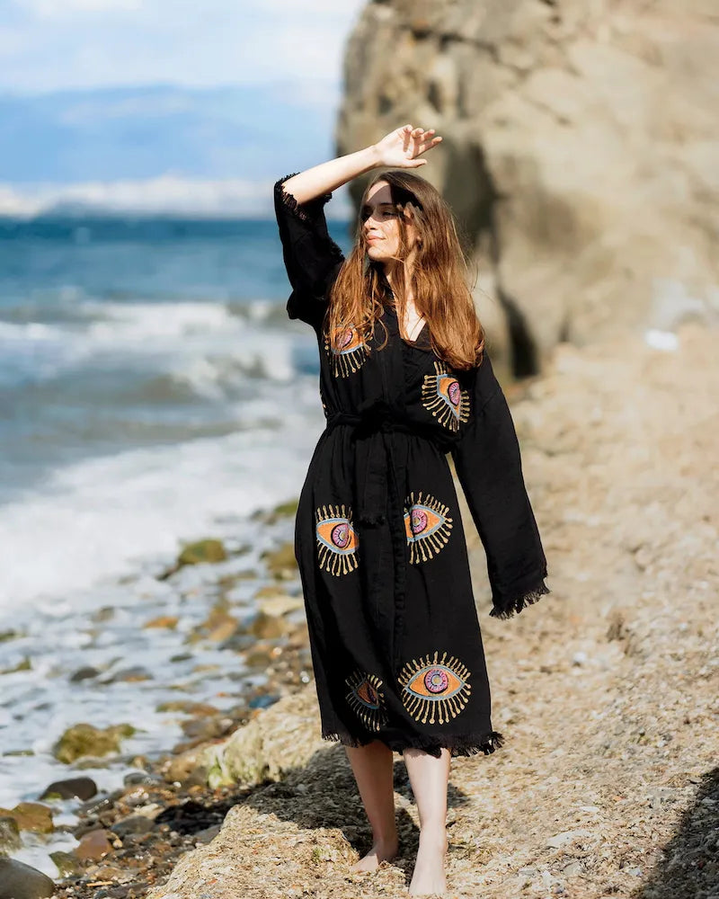 Woman in a black robe with eye designs standing on a rocky beach.