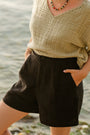 Woman in a beige top and black linen shorts by the water — European Flax linen, high-waisted pleated fit