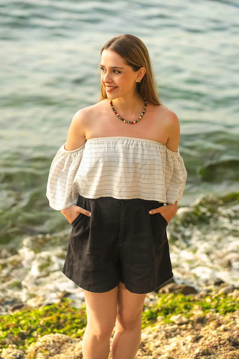 Woman wearing a white off-shoulder top and black linen shorts standing by a body of water.