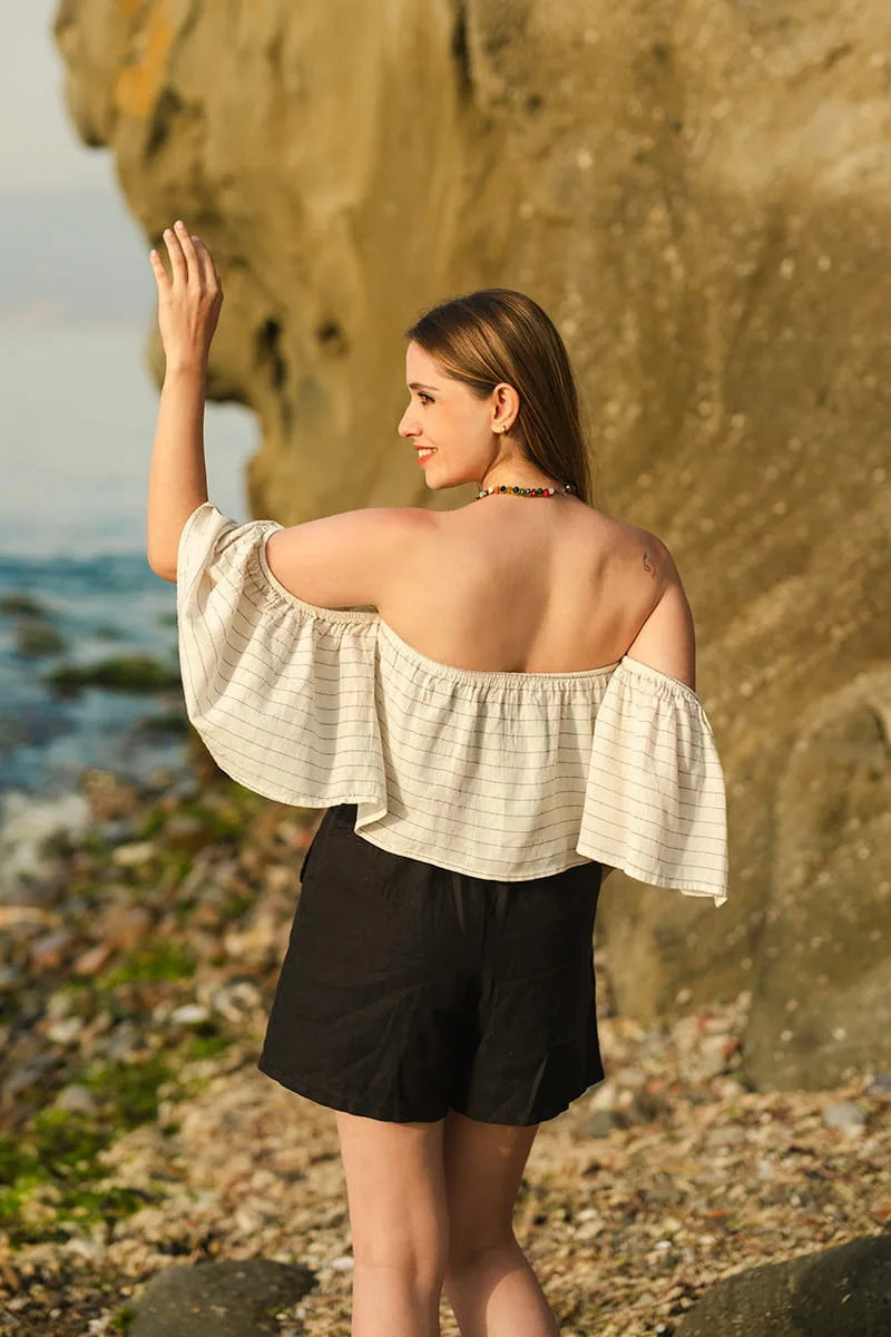 Woman in a white off-shoulder top and black linen shorts standing on a rocky beach.