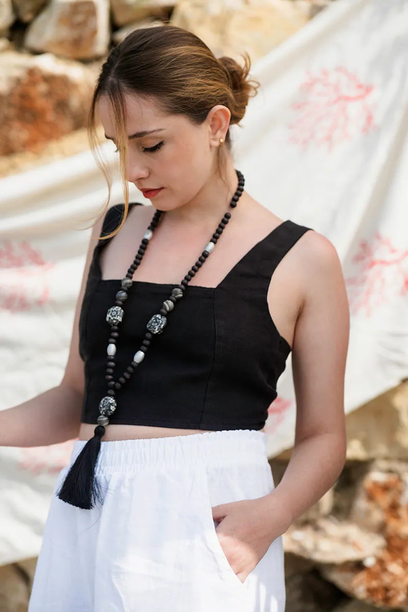 Woman wearing a black top and white pants with a long necklace, standing against a natural background.