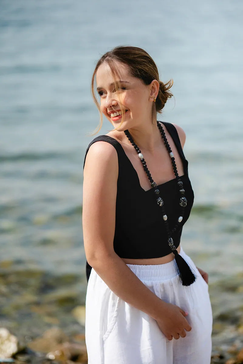 Woman wearing a black tie back top and white pants standing by a body of water.