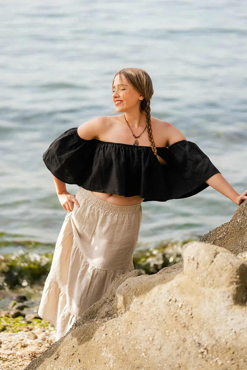 Woman in black off-shoulder top and beige skirt standing on rocks by a body of water.