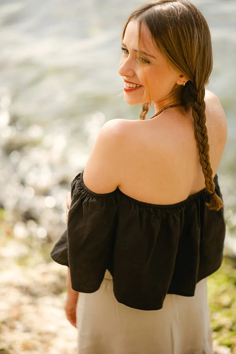 Woman wearing a black off-shoulder top with braided hair, standing by a body of water.