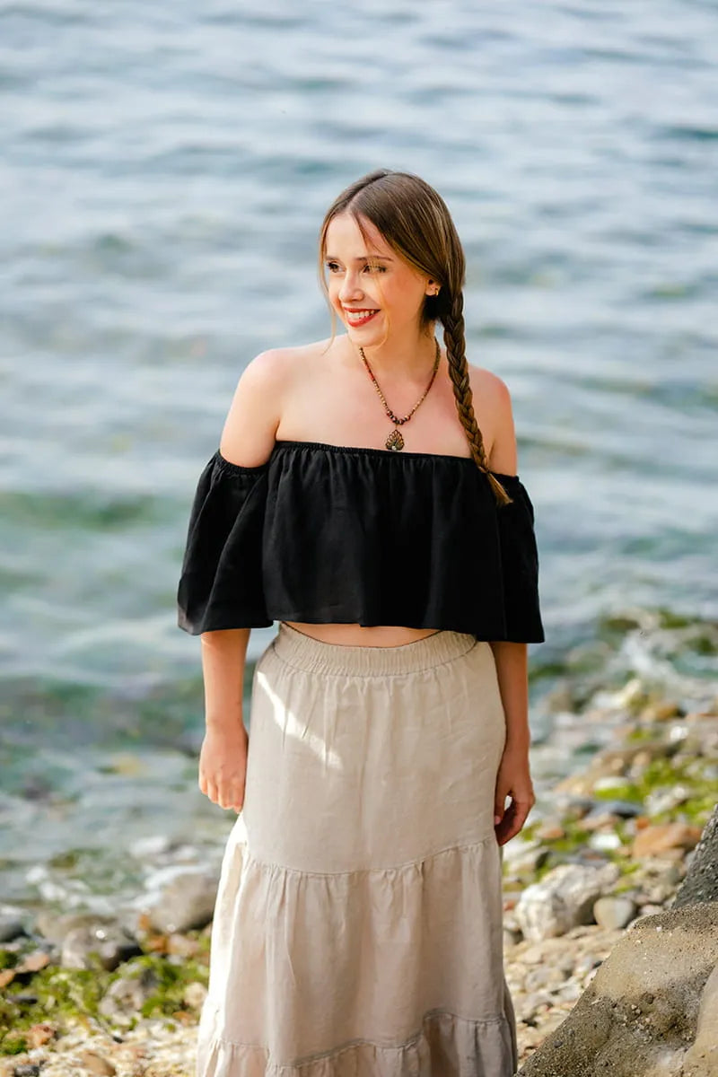 Woman standing by a body of water wearing a black off the shoulder crop  top and beige skirt.