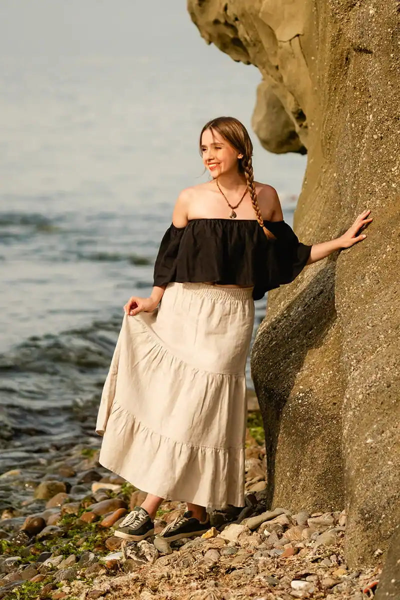 Woman in black off the shoulder top and beige skirt standing by a rocky shore with water and sky in the background