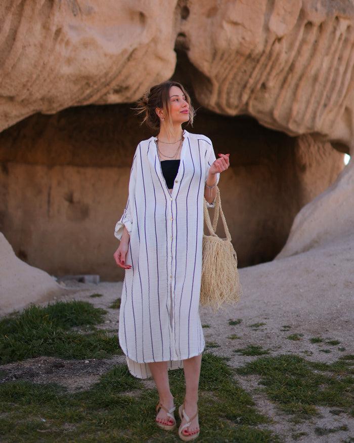 A woman standing in a natural archway wearing a white and black striped midi shirt dress with long sleeves.