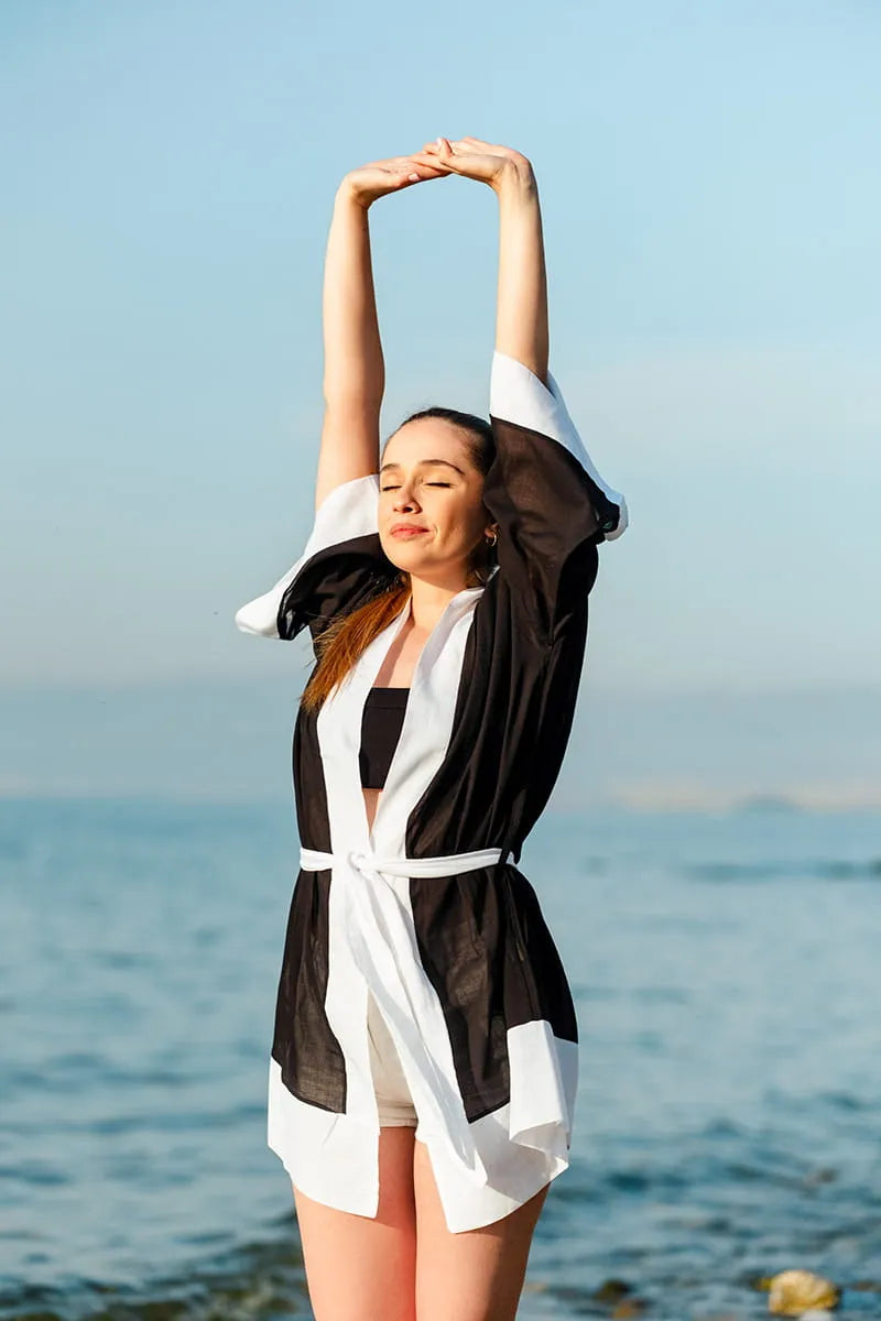 Woman with a Black Swimsuit Cover Up stretching outdoors with a clear blue sky and water in the background