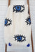 White blanket with black and blue evil eye patterns draped over a wooden ladder against a white brick wall.