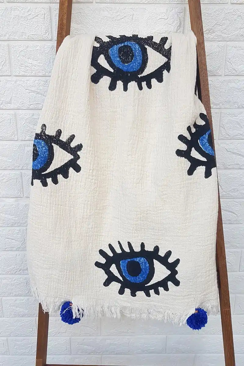 White blanket with black and blue evil eye patterns draped over a wooden ladder against a white brick wall.