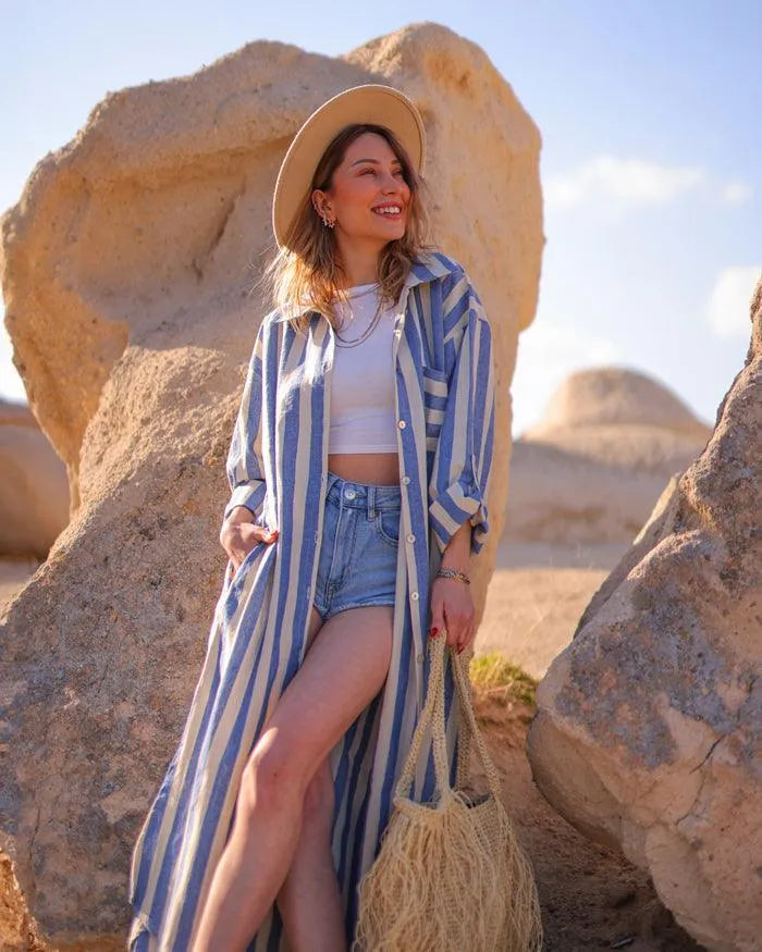 Woman in a striped shirt and shorts standing among large rocks with a straw hat and bag.