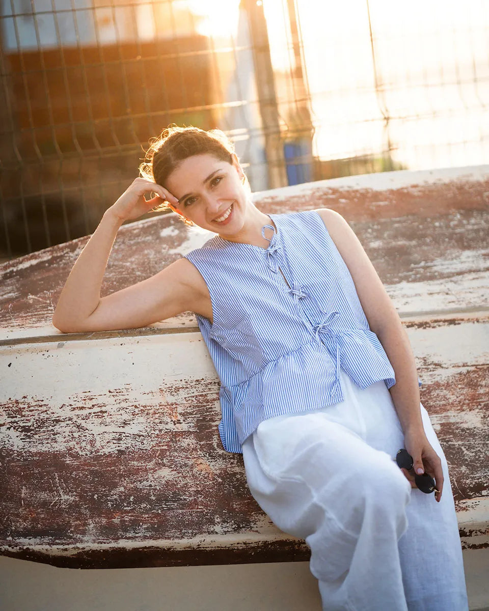 Woman in a blue striped top and white linenpants sitting on a wooden surface with a blurred background