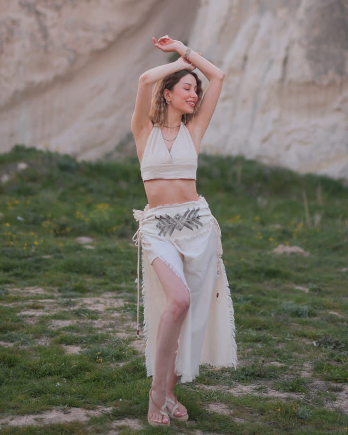 Woman in a beige top and Beige wrap skirt standing in a natural landscape with green grass and a rocky background.