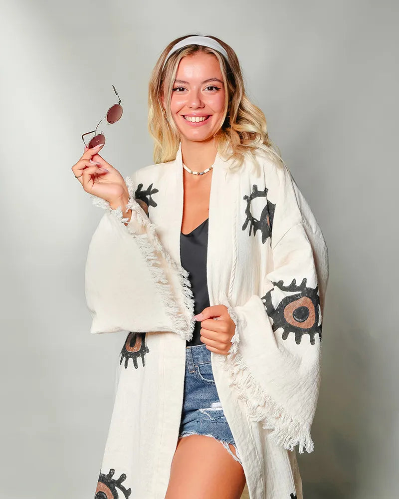 Woman wearing a white cardigan with brown evil eye patterns, holding sunglasses, on a plain background
