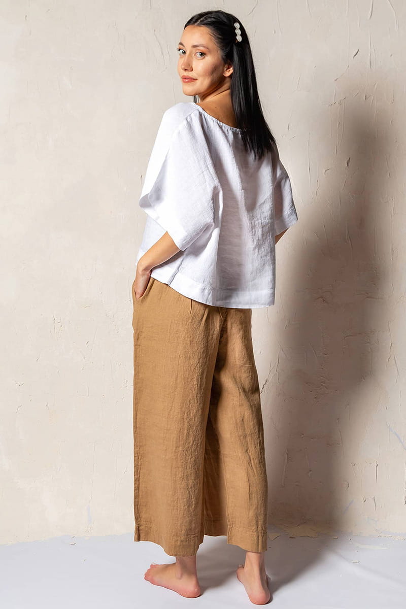 Woman wearing a white top and brown linen pants against a plain wall.
