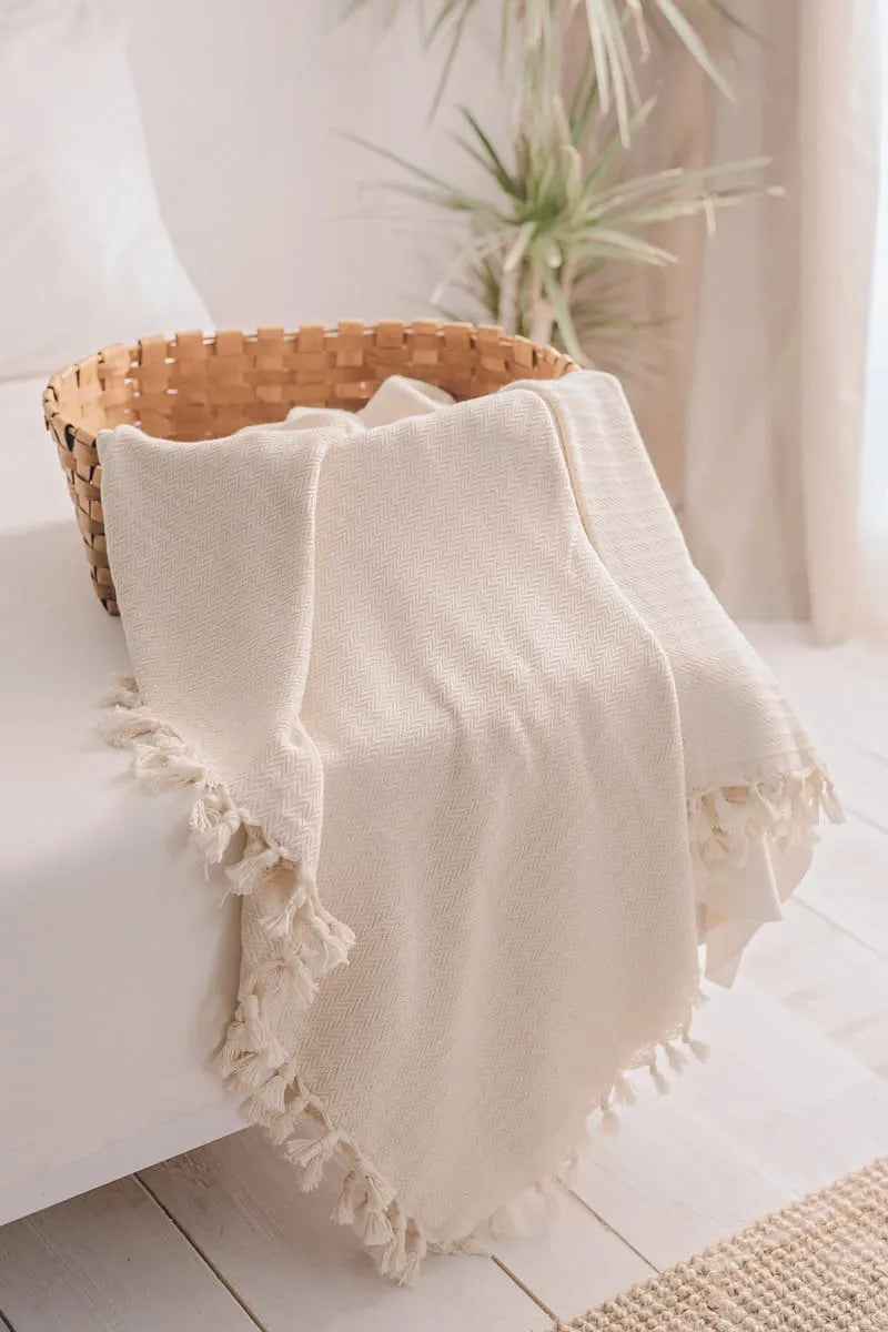 cream blanket with tassels draped over a white surface, with a wicker basket and plant in the background.